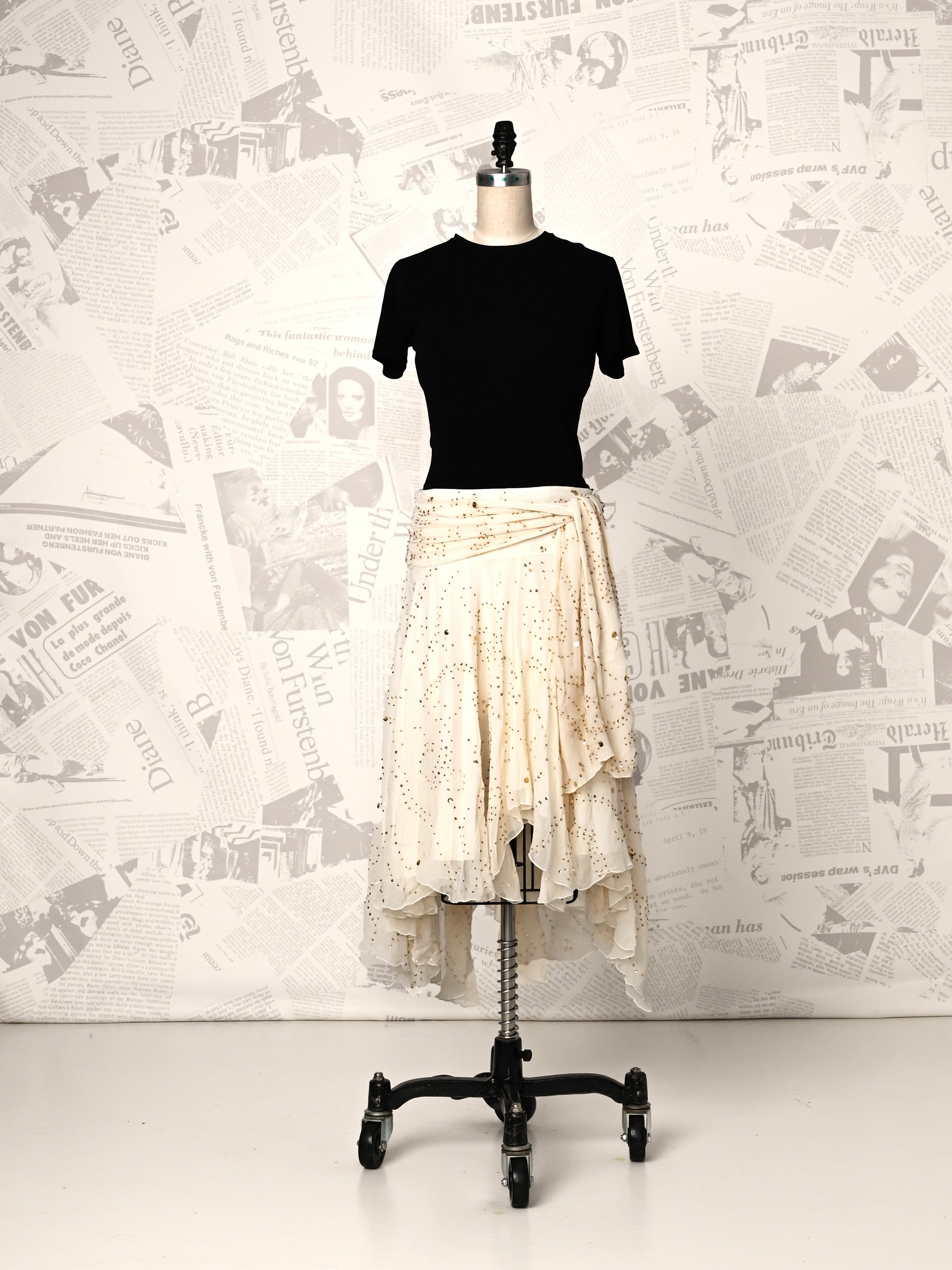 Mannequin wearing a black top and beige skirt against a newspaper-patterned wall.
