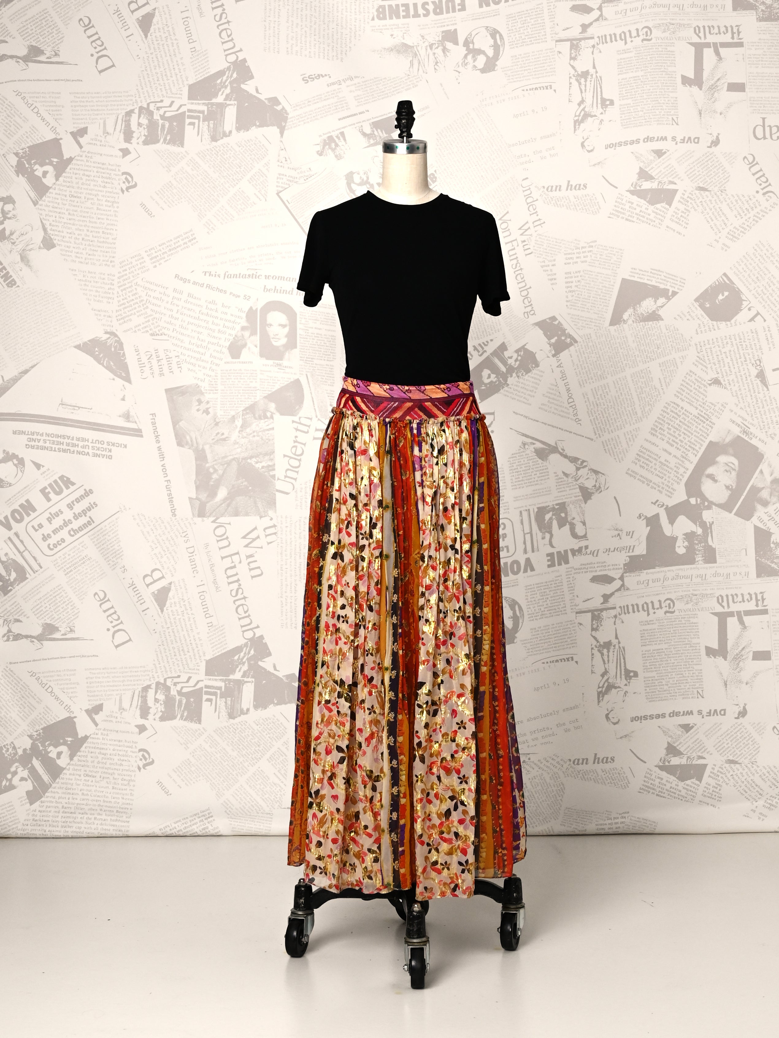 Mannequin wearing a black top and floral skirt against a newspaper-patterned wall.