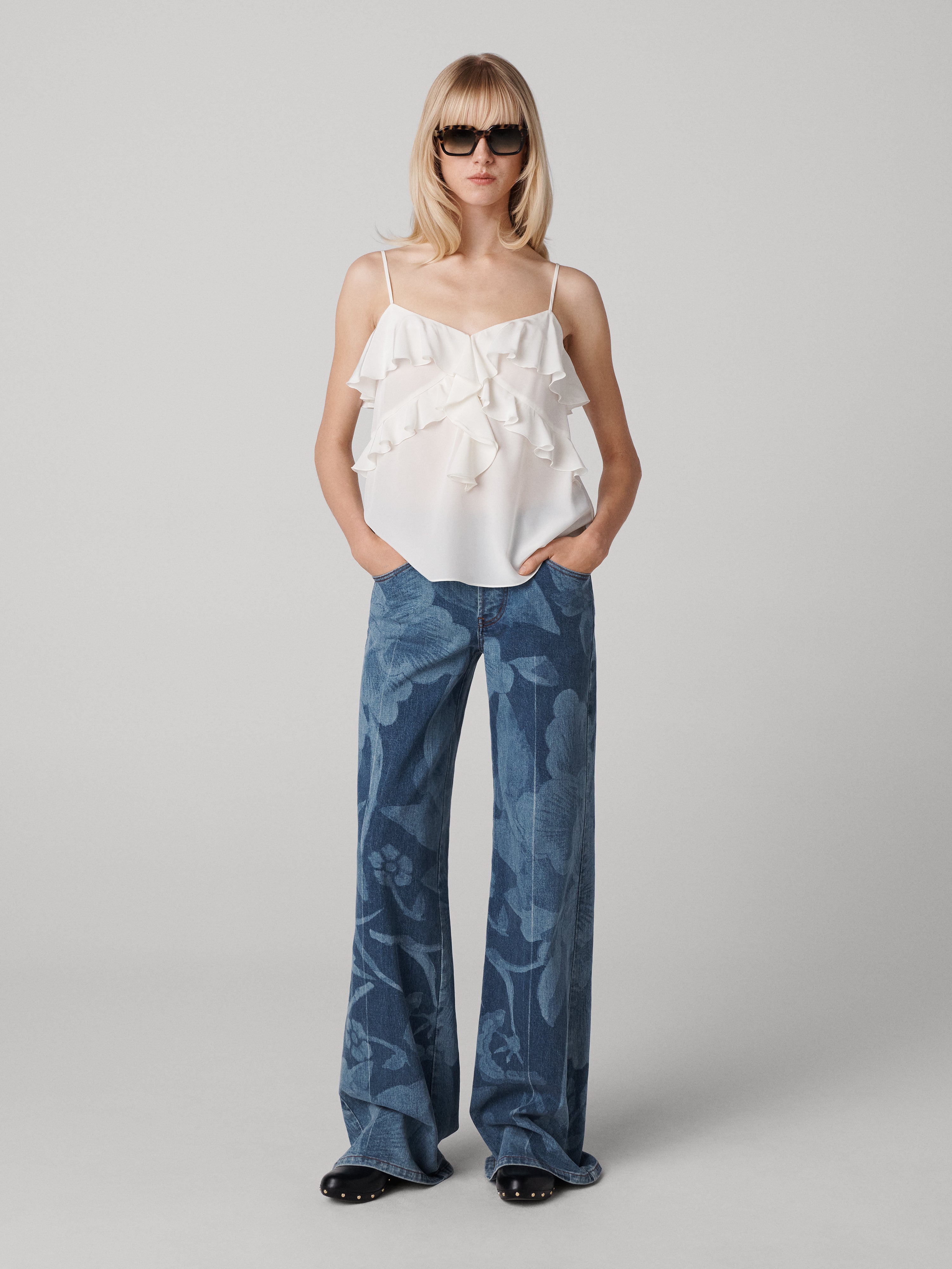 A woman wearing a white sleeveless top with ruffled neckline and blue denim pants with a floral print